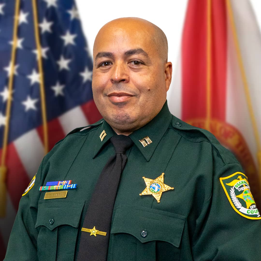 A formal, head-and-shoulders portrait of Captain Freddy Rivera of the Osceola County Sheriff's Office. He is a bald man with a warm smile, wearing a dark green law enforcement uniform jacket, a black tie with a gold star tie tack, and a gold star badge pinned to his chest. His collar features gold captain rank insignia, and a nameplate and ribbon bars are pinned above his right pocket. On his left sleeve is the Osceola County Sheriff's Office patch. The background features the blurred American flag on the left and a state flag on the right.