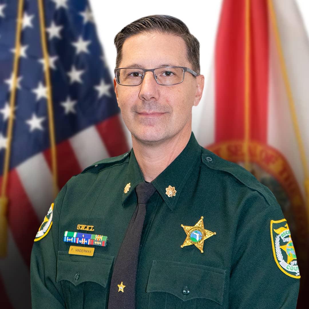 Official headshot of Major F. Hinderman wearing a dark green law enforcement uniform with a gold five-point star badge, service ribbons, and a nameplate. He is wearing glasses and is positioned between the United States flag and the Florida state flag.